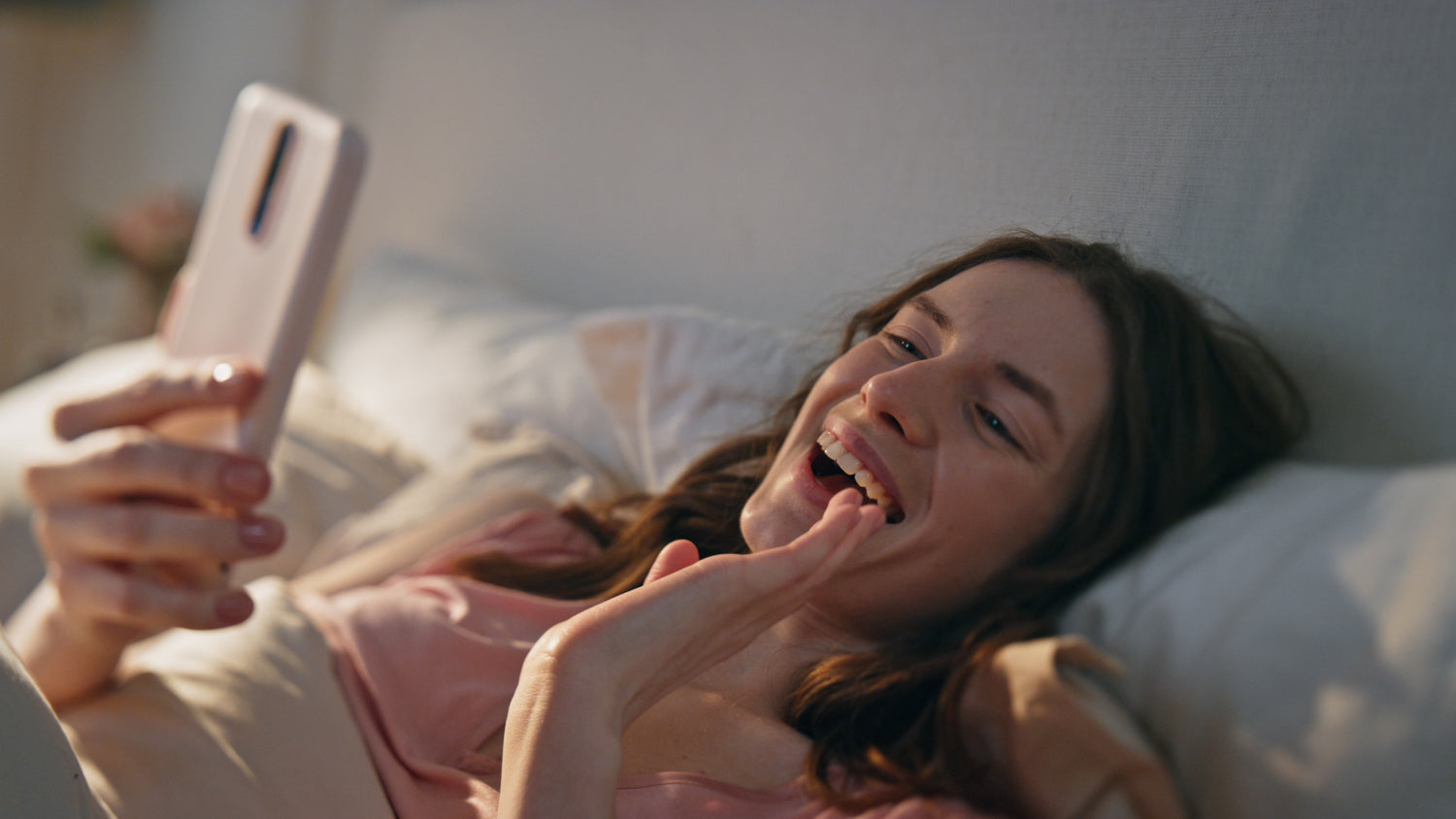Eine Frau die fröhlich lacht und im Bett liegt. Sie hält in der rechten Hand ihr Handy und mit der linken winkt sie dem Handy zu. Es handelt sich um einen Videoanruf.